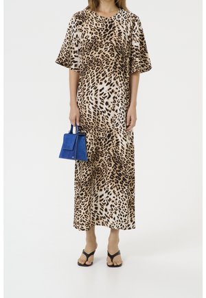 Femme portant une longue robe imprimé léopard, tenant un petit sac à main bleu et chaussée de sandales noires à brides sur fond blanc.