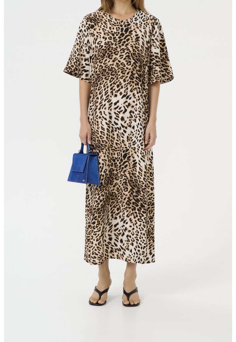 Femme portant une longue robe imprimé léopard, tenant un petit sac à main bleu et chaussée de sandales noires à brides sur fond blanc.