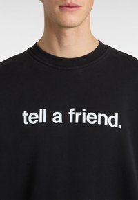 Sort sweatshirt med rund halsudskæring, prydet med fed hvid tekst, der står "fortæl en ven." Blødt stof med en glat overflade.