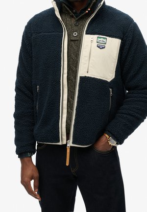 Mand iført navyblå fleecejakke med beige brystlomme og lynlås, mørk striktrøje under og mørke jeans med ur på venstre håndled.
