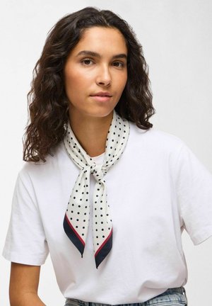 Kvinne med krøllete mørkt hår som har på seg en hvit t-skjorte og et hvitt skjerf med polkadotter bundet rundt halsen, vendt mot fronten.