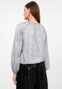 Hellgrau, strukturierte Bluse mit gerafftem Einsätzen und langen Ärmeln, kombiniert mit einem schwarzen Paillettenkleid, das einen Knopfverschluss hinten hat.