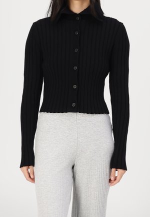 Femme portant un cardigan noir côtelé ajusté avec boutons et un pantalon en maille côtelée gris clair, debout devant un fond blanc uni.