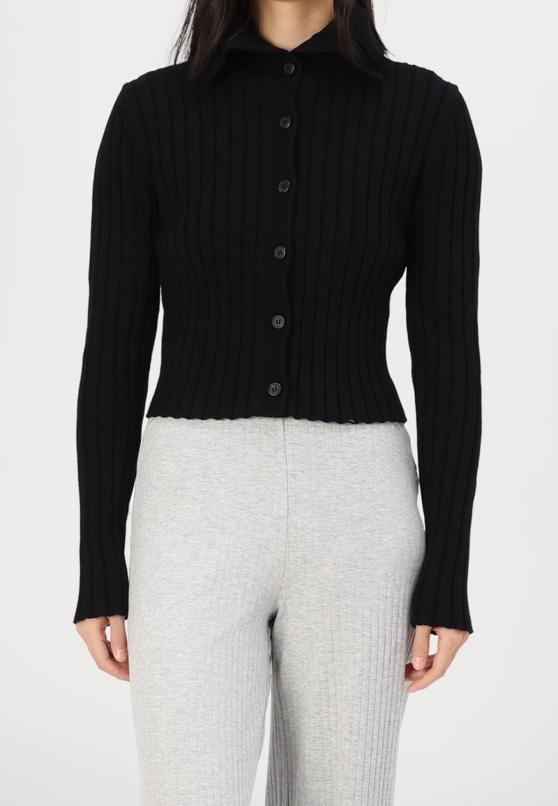Femme portant un cardigan noir côtelé ajusté avec boutons et un pantalon en maille côtelée gris clair, debout devant un fond blanc uni.