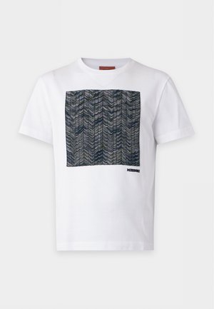 Man staat, draagt een wit T-shirt met korte mouwen met een groen patroon vierkant, beige broek en bruine instapschoenen tegen een witte achtergrond.