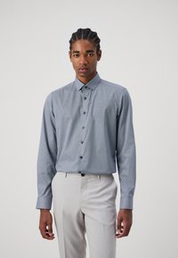Blaues Langarmhemd mit kleinem geometrischen Muster, knöpfbarem Kragen und dunklen Knöpfen, kombiniert mit hellen Hosen.