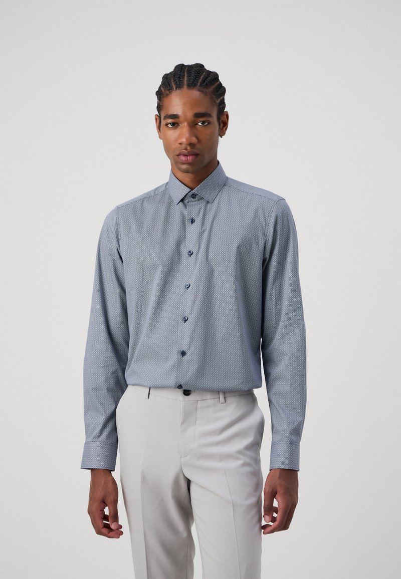 Blaues Langarmhemd mit kleinem geometrischen Muster, knöpfbarem Kragen und dunklen Knöpfen, kombiniert mit hellen Hosen.