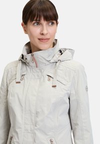Lichtgrijze waterdichte jas met capuchon, voorzien van gestikte accenten, twee voorzakken met rits en beige hardwaredetails.