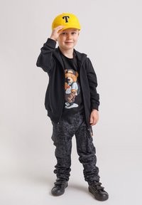 Czarna bluza z kapturem na zamek, t-shirt z grafiką niedźwiedzia, czarne spodnie o fakturze, żółta czapka z literą 'T' oraz czarne buty na paski.