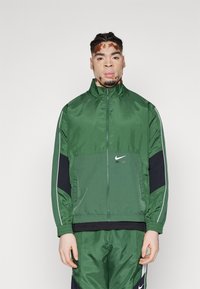 Chaqueta de chándal verde de Nike con cremallera completa, cuello alto y paneles laterales en color marino. Presenta un brillo sutil y detalles en piping blanco.