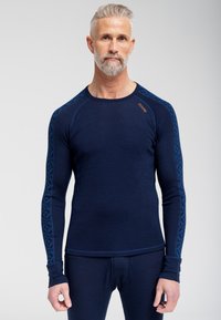 Marineblå langærmet termotrøje lavet af let stof, med et geometrisk mønster i en mørkere blå farve langs ærmerne og små orange mærker.