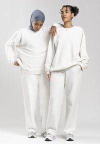 Zwei Frauen tragen passende weiße Sweatshirts und Hosen, eine mit einem grauen Hijab, stehen vor einem schlicht hellen Hintergrund.