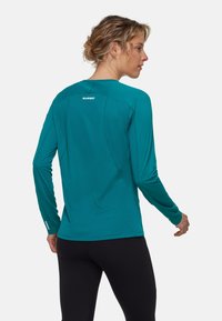 Langärmliges, türkisfarbenes Sportshirt aus strukturiertem Material, mit rundem Hals und Logo auf der Rückseite. Kombiniert mit schwarzen, eng anliegenden Leggings.