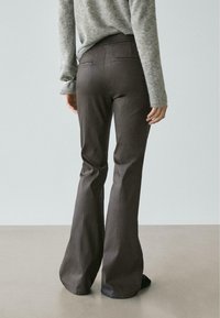 Personne portant un pantalon évasé gris foncé et un pull à manches longues gris clair, debout sur un sol neutre avec un fond clair.