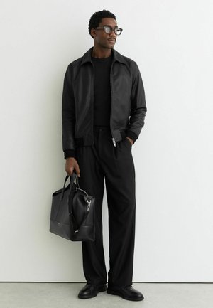 Hombre con chaqueta de cuero negra y pantalones de pierna ancha sosteniendo un bolso de cuero negro, de pie contra una pared blanca.