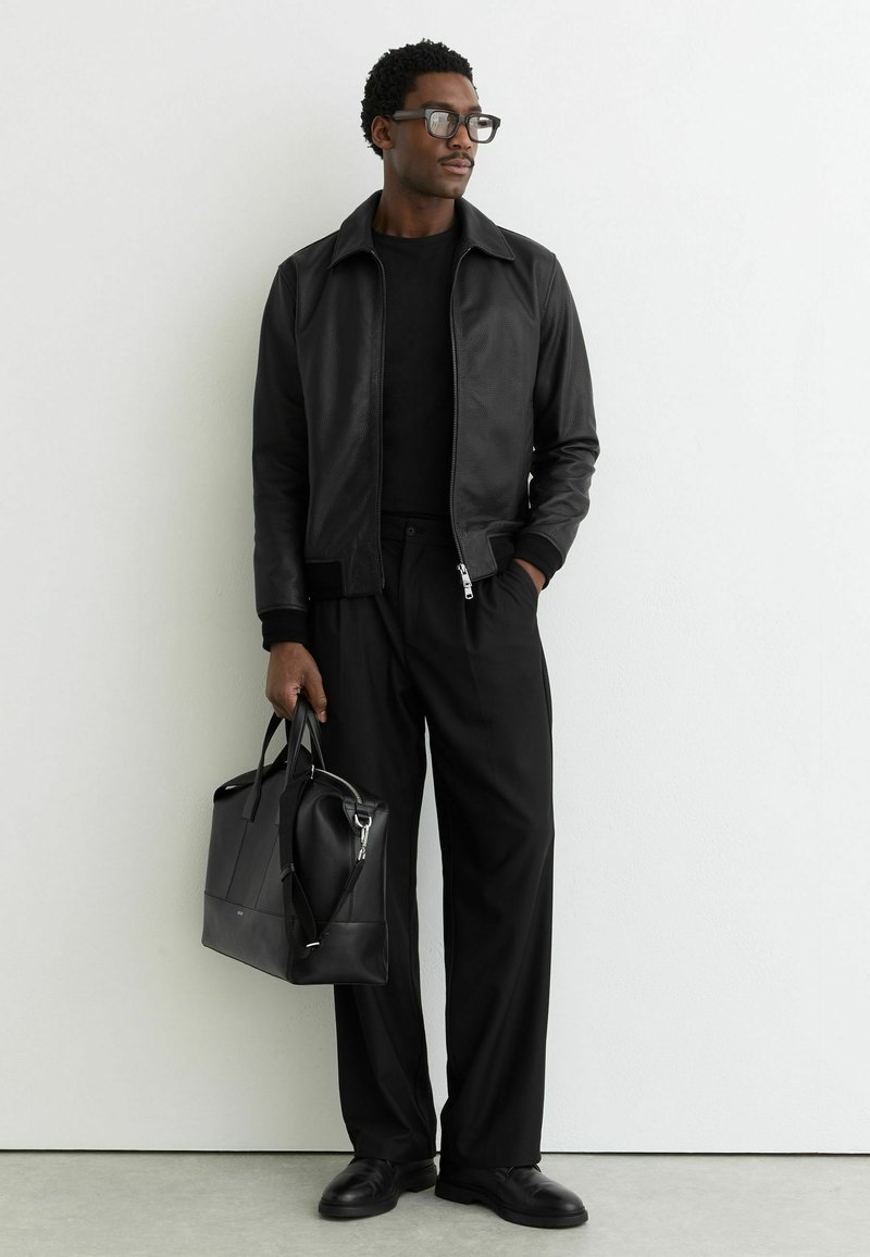 Mees mustas nahktagis ja laia säärega pükstega, musta nahast käekotti hoides, seisab valge seina ees.