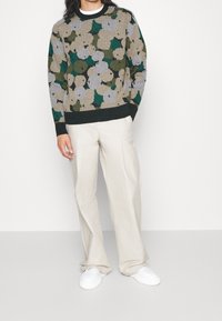 Beige, grüne und graue Strickpullover mit Blumenmuster; gerippter Halsausschnitt und Bündchen. Kombiniert mit hellbeigen Hosen und weißen Schuhen.