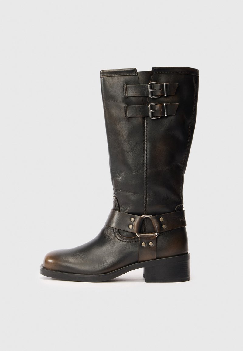 Botină din piele neagră până la jumătatea gambei, cu două catarame aproape de vârf și o curea cu detaliu în formă de inel metalic în jurul gleznei, pe un fundal alb.