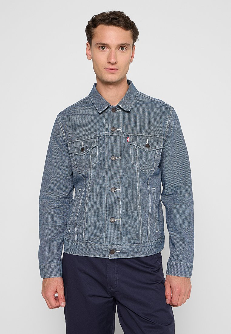 Veste en denim à motif rayé bleu avec un col, deux poches poitrine et fermeture à boutons sur le devant. Des détails de couture contrastés sont visibles.