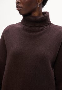Maglione lavorato a maglia marrone con collo alto a imbuto, caratterizzato da dettagli a coste sul collo e sui polsini, texture liscia e vestibilità rilassata.