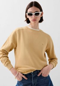 Gele katoenen sweatshirt met ronde hals en geribbelde manchetten, gecombineerd met donkere denimshorts. Witte zonnebril maakt de look compleet.