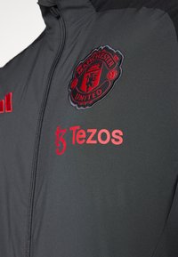 Chaqueta gris oscuro con cuello alto, que presenta el logotipo bordado de Manchester United en rojo y la marca Tezos en el lado izquierdo del pecho. Textura suave.