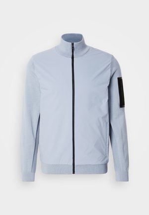 Heleheline jakk kõrge kraega, ees zipiga ja tekstuursete varrukatega. Omab musti aktsente vasakul käel ja soonikkoes mansetides.