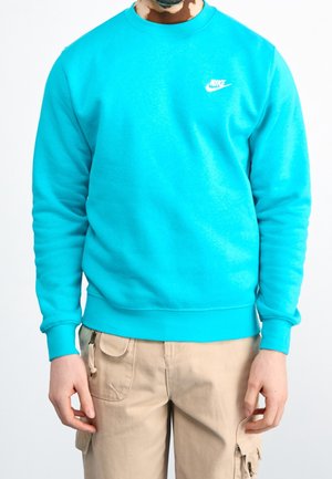 Homme portant un sweat-shirt Nike bleu vif associé à un pantalon cargo beige, debout devant un fond clair uni.