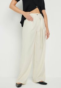 Pantalones de pierna ancha color crema, hechos de una tela suave, con cintura alta, frente plisado y un cinturón atado. Combinados con bailarinas negras.
