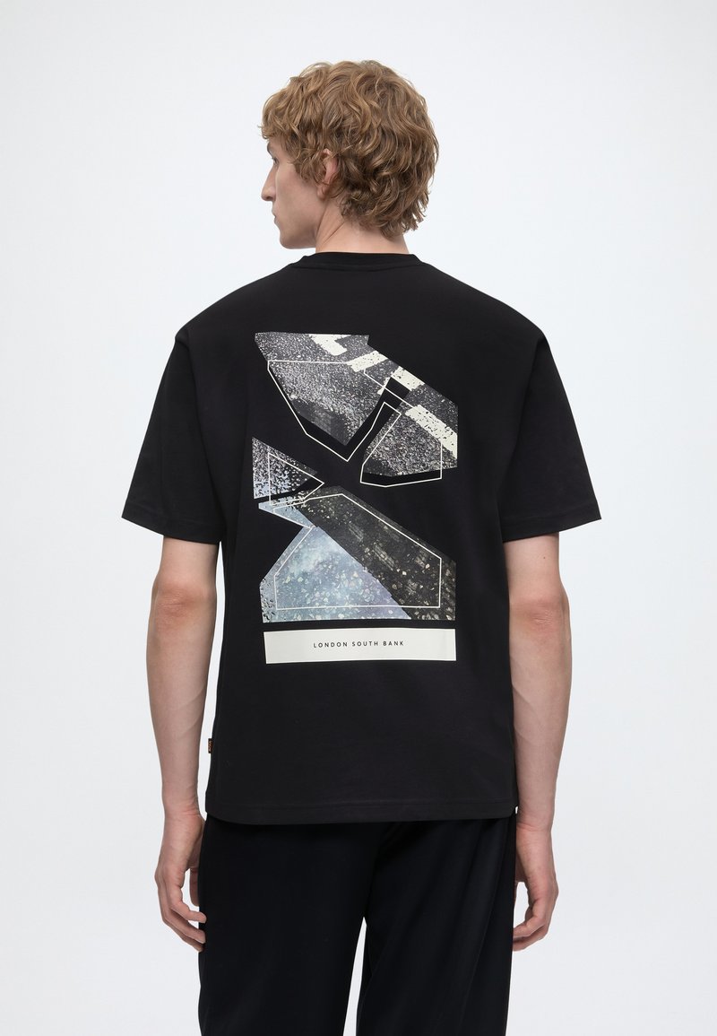Homme aux cheveux blonds bouclés portant un t-shirt noir avec un motif géométrique abstrait et le texte « London South Bank » dans le dos.