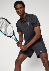 Black polo shirt with a logo and black shorts, holding a tennis racket with a blue accent, positioned for a tennis stance.