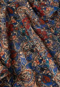 Tissu avec un motif complexe floral et paisley aux tons rouge, bleu, vert et beige, agrémenté de boutons noirs cousus le long des plis.