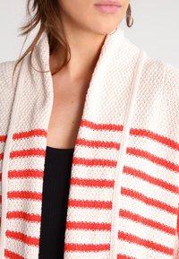 Krémový kardigan s texturovaným materiálem, zdobený červenými vodorovnými pruhy, s otevřeným předním dílem a uvolněným střihem, který zdůrazňuje pohodlný design.