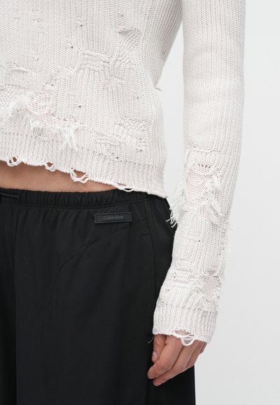 Pulover alb tricotat cu margini uzate și modele, asortat cu pantaloni negri conici, având o etichetă Calvin Klein pe talie.