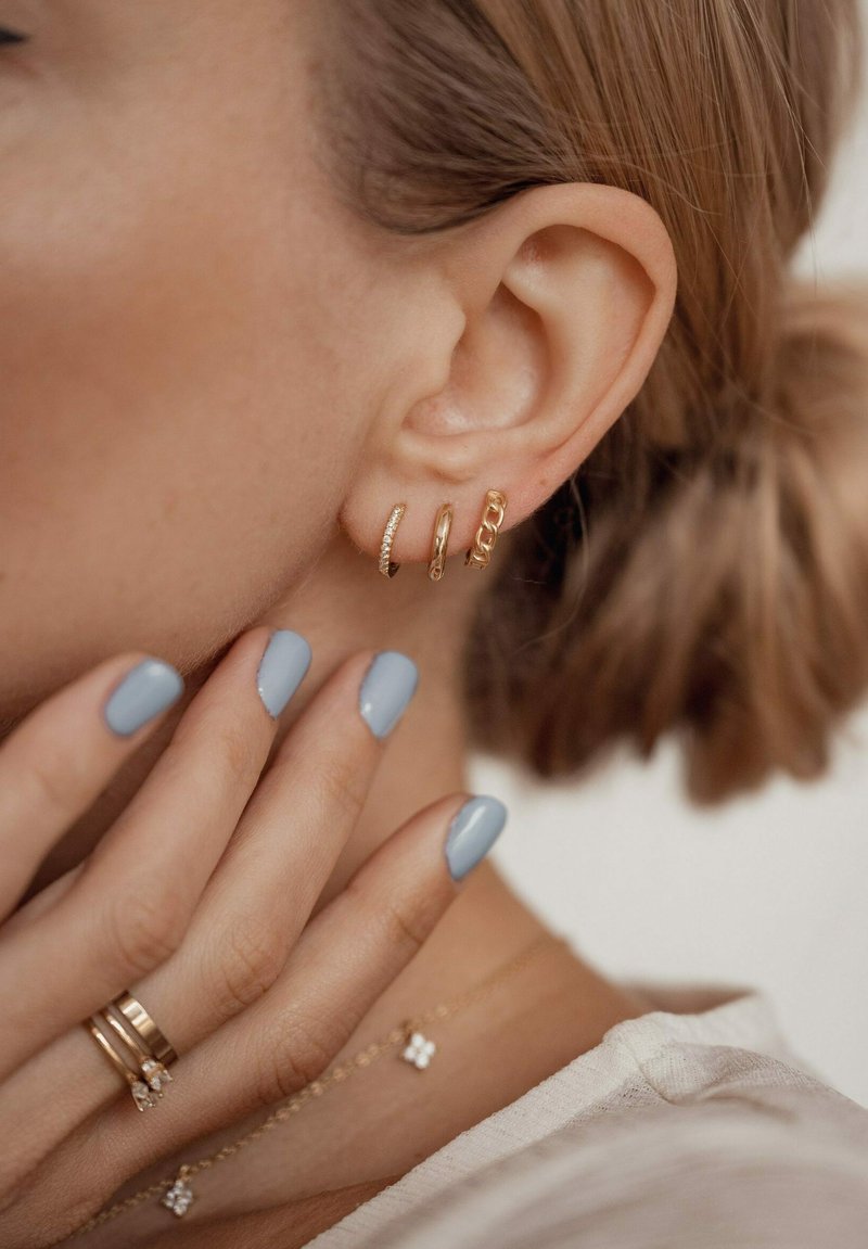 Pendientes de aro dorados en tres estilos: uno liso, uno texturizado con pequeñas piedras y uno con diseño de eslabones de cadena. Uñas pintadas de azul claro.