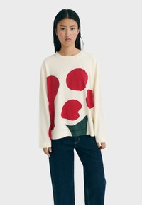Long-sleeve beige top with large red and green abstract shapes, crew neckline, relaxed fit, and soft fabric. Worn with dark denim pants.