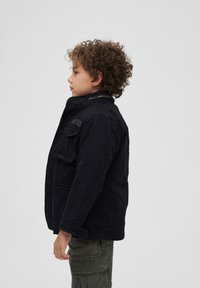 Juodas lauko stiliaus džemperis su aukštu apykakle, priekinėmis kišenėmis ir užtrauktuku, dėvimas vaiko prieš neutralią fono aplinką.