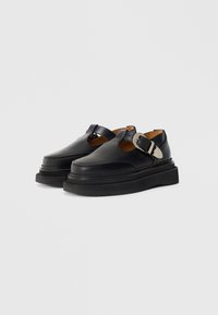 Zwarte leren schoenen met een rond neu en een dikke plateauzool. Voorzien van een verstelbare gesp over de wreef en zichtbare stiksels.