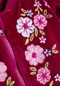 Giaccone borgogna con zip, caratterizzato da ricami floreali in bianco, rosa e blu con foglie verdi, realizzato in un tessuto morbido e testurizzato.