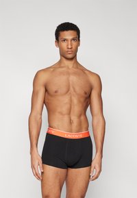 Svarta boxerkalsonger med en röd "LACOSTE" midja. Tyget verkar glatt med en åtsittande design och minimala sömmar.