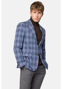 Niebieska kratkowana marynarka z brązowymi akcentami, wykonana z teksturowanej tkaniny, z zapięciem na jeden guzik i dwoma kieszeniami z klapkami. Nosi się ją na brązowym golfie.