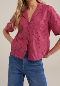 Kortärmad, knappad skjorta i strukturerad magenta tyg med broderade blommönster och utskärningar, tillsammans med blå jeans.