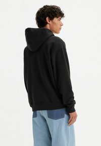 Black hoodie with a kangaroo pocket, ribbed cuffs, and a drawstring hood. Worn with light blue relaxed-fit jeans featuring patch details.