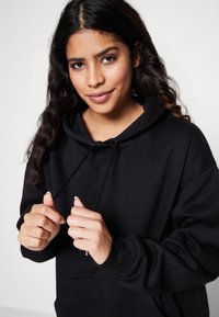 Schwarzer Kapuzenpullover mit Kängurutasche, ausgestattet mit Kordeln und gerippten Bündchen. Weicher Stoff, lässige Passform, sichtbare Nähte.