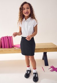 Witte kortärmige shirt met een zak en een zwarte rok. Zwarte gepatineerde leren schoenen met een gesp en sokken met rode accenten. Roze boeken op de achtergrond.