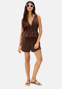 Brun peplum jumpsuit i lättviktig tyg, med djup V-ringning, justerbar midjeband och fransad fåll. Bärs med platta sandaler.