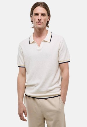 Jeune homme aux cheveux bruns mi-longs, portant un polo blanc avec un bord foncé et un pantalon beige, debout avec une main dans la poche.