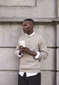Pulover bej cu dungi, cu guler rotund, purtat peste o cămașă albă. Ținând o cană de cafea de unică folosință, stând în fața unui perete texturat.