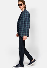Marynarka w kratę w ciemnozielonym i granatowym kolorze, o dopasowanym kroju, zestawiona z ciemnymi dżinsami i granatowymi trampkami. Model nosi czarne okulary przeciwsłoneczne, zwrócony w lewo.