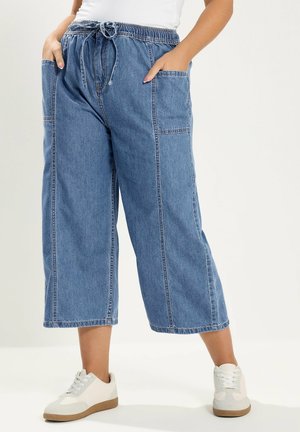 Losse blauwe denim broek met korte pijpen, grote voorzakken en een taille met trekkoord, gecombineerd met witte sneakers en een witte top.